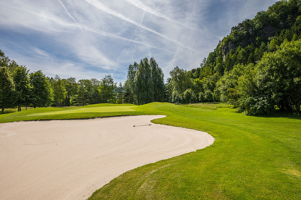 Ein heller Golfplatz mit einem Sandbunker auf der linken Seite, saftig grünem Gras und umliegenden Bäumen unter einem teilweise bewölkten Himmel, der eine heitere Atmosphäre hervorruft.