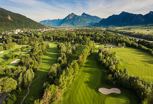 Luftaufnahme des Golfplatzes vor der Kulisse majestätischer Berge unter einem klaren blauen Himmel. Die Szene ist heiter und malerisch.