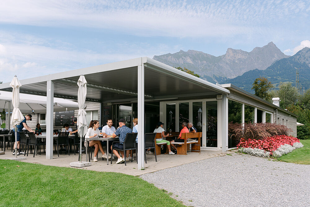 Unser Golf Bistro, in dem Gäste unter einer schattigen Terrasse speisen. Umgeben von Grünflächen und Bergen vermittelt die Szenerie eine entspannte und malerische Atmosphäre.