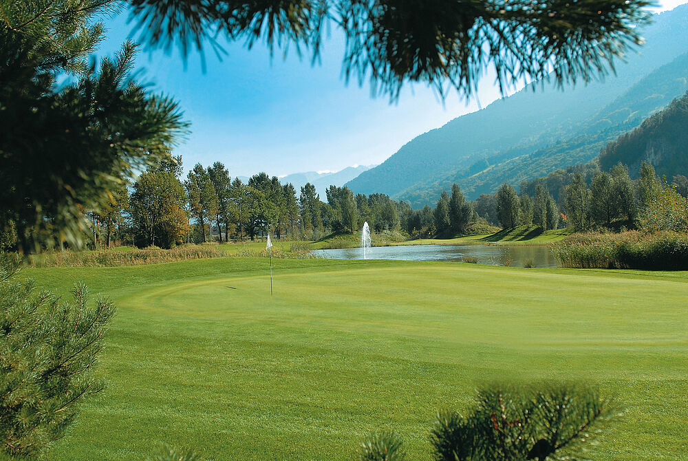 Ein Golfplatz, umrahmt von Bäumen, mit einem ruhigen Teich und einem Springbrunnen in der Ferne, vor der Kulisse der Berge und unter einem klaren blauen Himmel.